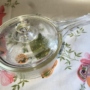 May include: A clear glass saucepan with a matching lid, featuring an etched floral pattern on the lid and a long, ribbed handle. The glass is transparent, showing the intricate decorative details on the surface of the lid and the handle.