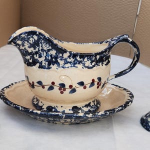 Può includere: Una salsiera in ceramica con piattino integrato e due candelabri abbinati. I pezzi sono color crema con un motivo a spugnatura blu scuro e un decoro dipinto a mano raffigurante tralci di vite con piccole bacche rosse e foglie blu.