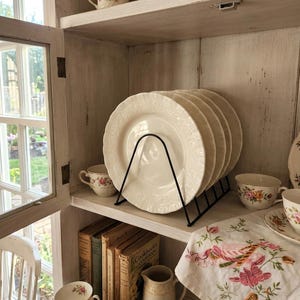 May include: A set of four cream-coloured ceramic plates with embossed floral rims, held upright in a black wire metal rack. The plates are displayed on a shelf alongside floral teacups, a small pitcher, and several vintage books.