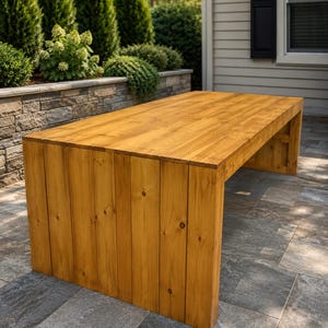 May include: A rectangular wooden table featuring a light brown finish with visible wood grain patterns. The table is constructed from vertical planks that form both the flat top surface and the two solid side supports, creating a continuous design.