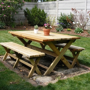 Könnte beinhalten: Ein hölzerner Picknicktisch mit angebrachten Bänken. Der Tisch und die Bänke sind aus hellem Holz gefertigt und haben ein X-förmiges Beindesign. Ein Blumentopf steht auf dem Tisch. Der Tisch ist von grünem Gras und einem Ziegelrand umgeben.