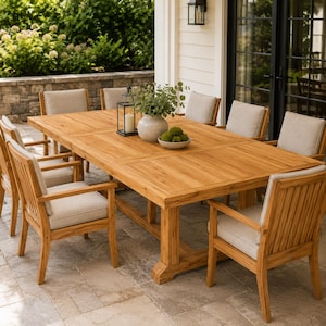 May include: A rectangular teak wood dining table with a trestle base and eight matching chairs featuring vertical slat backs and light beige upholstered seat cushions. A black metal lantern and a ceramic bowl with green spheres sit on the table.