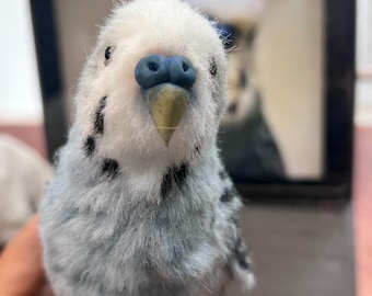 Puede incluir: Una figura artesanal de periquito, difusa, de color azul claro y blanco con una apariencia texturizada y realista. El ave presenta una cabeza blanca, un pecho azul claro con marcas negras, una cera azul prominente y un pico puntiagudo de color amarillo pálido.