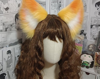 May include: A pair of large, pointed faux fur animal ears in a gradient of orange, yellow, and white, attached to a headband. The ears are positioned on a mannequin head featuring long, wavy brown hair with bangs, creating a realistic look.
