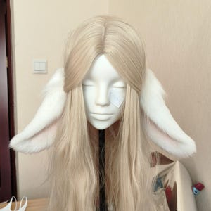 May include: A long, straight, pale blonde wig styled with a center part, featuring two large, plush, white animal-style ears attached to the sides. The ears have a soft, furry texture with light pink inner detailing, positioned symmetrically on the head.