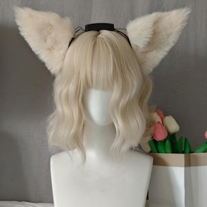 Op de afbeelding: Een wit etalagehoofd met een halflange, golvende blonde pruik met pony en een paar grote, crèmekleurige imitatiebont dierenoren bevestigd aan een dunne zwarte haarband met een klein rechthoekig elektronisch onderdeel.