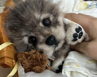 Puede incluir: Un pequeño cachorro de peluche realista con pelaje gris y blanco, ojos oscuros y nariz negra. El cachorro tiene almohadillas negras visibles en las patas y sostiene un pequeño oso de peluche marrón sobre una tela blanca con lunas y estrellas.