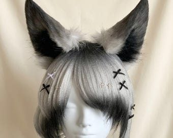 Puede incluir: Una peluca de cabello liso y en capas con un degradado de negro a blanco plateado, que presenta grandes orejas de animal puntiagudas de piel sintética en gris y negro. El cabello está adornado con pequeñas cruces de cinta negra y blanca y dijes de cruz plateados.