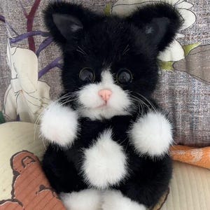May include: A plush toy cat with black fur and white patches on its face, chest, and paws. The cat has large, dark, round eyes, a small pink nose, and long white whiskers. It is depicted in a seated position with its front paws raised slightly.