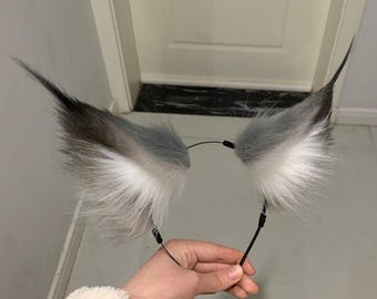 Puede incluir: Una diadema con dos orejas de animal puntiagudas de piel sintética. Las orejas tienen un patrón de color degradado, que va del gris oscuro en las puntas al gris claro en el centro y blanco en la base. La diadema es fina y negra.