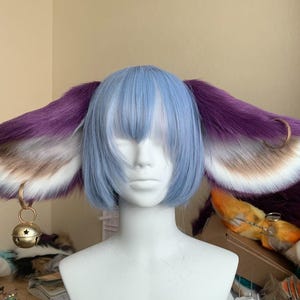 Könnte beinhalten: Eine hellblaue Bob-Perücke auf einem weißen Schaufensterpuppenkopf mit großen, flauschigen Tierohren aus violettem und weißem Kunstfell an den Seiten. An jedem Ohr befindet sich ein goldfarbener Metallring mit einer hängenden Glocke.