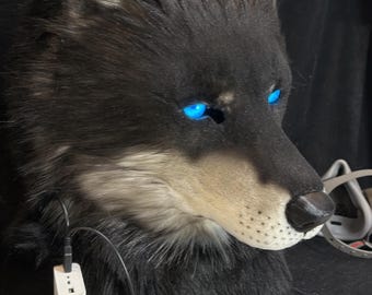 Puede incluir: Una máscara de cabeza de lobo realista con piel sintética negra y gris, orejas puntiagudas con puntas blancas y ojos LED azules brillantes. La máscara incluye una nariz negra, un hocico blanco y un cable negro conectado a una batería portátil.