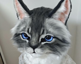 Puede incluir: Una máscara de cabeza de gato realista con piel sintética gris y blanca, orejas puntiagudas con puntas oscuras y llamativos ojos azules brillantes. La máscara está adornada con una prenda de encaje blanco drapeada alrededor del cuello.