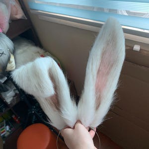 May include: A pair of tall, upright rabbit ears attached to a thin metal headband. The ears are covered in long, white faux fur with subtle brown and pinkish-tan accents along the inner edges and base, creating a realistic, textured appearance.
