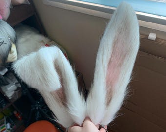 Puede incluir: Un par de orejas de conejo largas y erguidas unidas a una diadema de metal fina. Las orejas están cubiertas de piel sintética blanca de pelo largo con puntas marrones y un forro interior rosa suave, con una oreja recta y otra doblada.