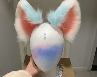 Puede incluir: Una diadema con dos orejas de animal grandes y erguidas hechas de piel sintética larga y esponjosa. Las orejas son blancas en los bordes exteriores, pasando a un azul pastel suave en las puntas, con un interior rosa vibrante que contrasta.