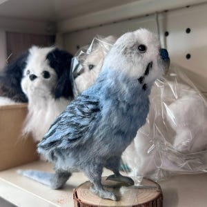 Puede incluir: Una figura artesanal y esponjosa de un periquito azul y blanco con marcas negras en las alas, sobre una base circular de rodaja de madera. El p&aacute;jaro tiene un peque&ntilde;o pico azul y ojos oscuros, frente a un juguete de peluche blanco visible.