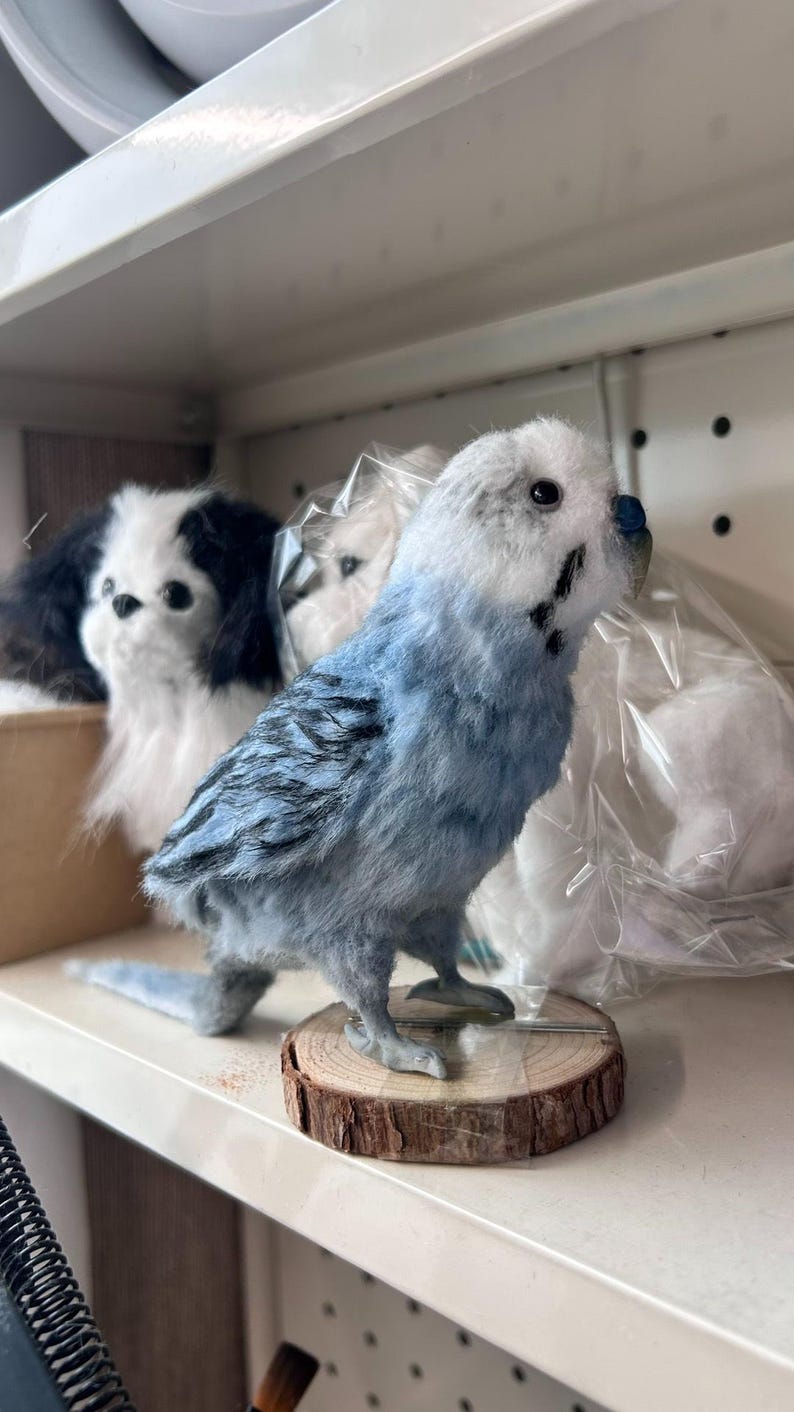 Puede incluir: Una figura artesanal y esponjosa de un periquito azul y blanco con marcas negras en las alas, sobre una base circular de rodaja de madera. El p&aacute;jaro tiene un peque&ntilde;o pico azul y ojos oscuros, frente a un juguete de peluche blanco visible.