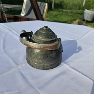 May include: A vintage, antique metal teapot with a rounded body and a domed lid. The teapot has a handle and a spout. The metal has a weathered, aged appearance with some rust-colored accents. The teapot is sitting on a white tablecloth.