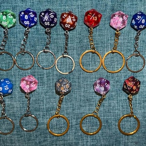 May include: A collection of fourteen twenty-sided polyhedral dice keychains arranged in two rows. Each die features white numbers and is attached to a metal chain and ring. The dice come in various colours including white, green, blue, purple, red, pink, and orange.