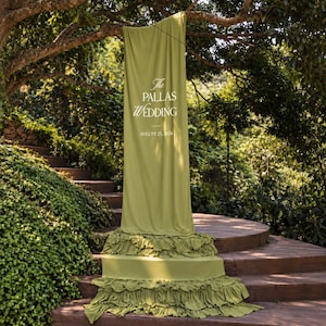 Puede incluir: Una larga pancarta de tela verde oliva con texto impreso en blanco que dice The PALLAS WEDDING y AUGUST 25, 2026. La pancarta cuelga de una rama de árbol y presenta múltiples capas de volantes fruncidos en la parte inferior de la tela.