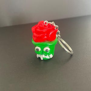 Peut inclure: Un porte-clés représentant une rose rouge vif sortant d'un pot vert ressemblant à un monstre. Le pot possède deux grands yeux blancs à pupilles noires et une rangée de dents blanches pointues sur le bord inférieur. Un anneau en métal argenté est fixé.