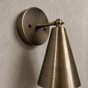 May include: A wall-mounted sconce light fixture featuring a circular backplate and a conical shade, both finished in a brushed antique brass tone. The lamp includes a short horizontal arm with a swivel joint connecting to the downward-facing shade.