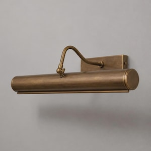 May include: A wall-mounted picture light fixture with an antique brass finish. The design features a rectangular backplate, a curved adjustable arm with a small tightening knob, and a long cylindrical light housing with a smooth metallic surface.
