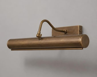 May include: A wall-mounted picture light fixture with an antique brass finish. The design features a rectangular backplate, a curved adjustable arm with a small tightening knob, and a long cylindrical light housing with a smooth metallic surface.