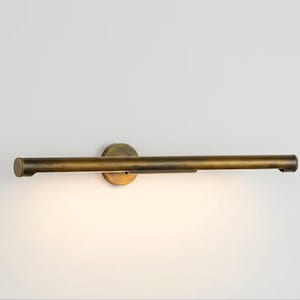 Op de afbeelding: Een horizontale wandlamp met een antieke messing afwerking. Het ontwerp bestaat uit een lange, cilindrische buis bevestigd aan een ronde basisplaat, die een zachte, warme gloed uitstraalt vanaf de onderkant van de metalen behuizing.