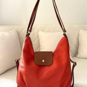 Pode incluir: Uma vibrante bolsa de ombro de couro laranja com alças de couro marrom e fecho de aba. A bolsa tem um zíper na parte superior e uma pequena aba marrom com um botão. A bolsa é segurada por uma mão.