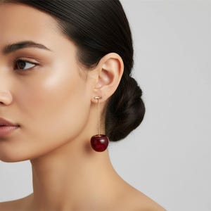 May include: A drop earring featuring a thin gold-toned metal bar attached to a single, smooth, deep red cherry-shaped charm. The earring hangs vertically from the earlobe, showcasing a minimalist design with a polished finish on the cherry.