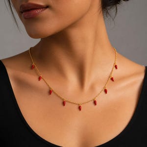 May include: A gold chain necklace with small, red chilli pepper pendants. The necklace is worn against a black top. The chilli peppers are evenly spaced along the chain and have a glossy finish.