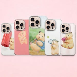 May include: Five phone cases with watercolor illustrations of hamsters. Each case features a different design, including a hamster with a red object, a hamster with a heart, a hamster in a blue vest, a hamster with a tennis racket, and a hamster with a pink object.