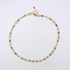 May include: A beaded necklace featuring a repeating pattern of translucent peach-colored round beads, small faceted dark brown beads, and tiny green beads. The necklace includes a gold-toned metal lobster clasp and an adjustable chain extender.