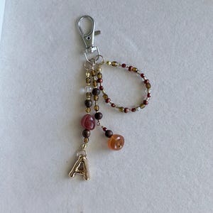 May include: A keychain featuring a silver-toned lobster clasp attached to a series of beads in shades of brown, clear, and deep red. The design includes a small loop of beads and a dangling gold-toned letter A charm at the end of a beaded strand.