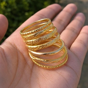 May include: A stack of six gold-toned metal rings, each featuring distinct surface textures including smooth bands, intricate floral engravings, and serrated edges, arranged in a cascading formation to display their varied metallic finishes.