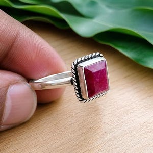 May include: A silver ring featuring a rectangular, faceted deep pink gemstone set in a bezel with a twisted rope-style border. The band is smooth and polished, providing a clean contrast to the textured setting surrounding the central stone.