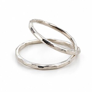 Könnte beinhalten: Zwei silberne Ringe mit gehämmerten Texturen, einer liegt auf dem anderen. Die Ringe sind kreisförmig und haben eine leicht unregelmäßige Form, die das Licht reflektiert. Der Hintergrund ist schlicht weiß und hebt den Schmuck hervor.