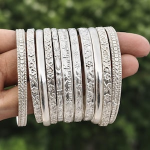 Op de afbeelding: Een set van tien zilverkleurige metalen armbanden met diverse reliëfpatronen, waaronder bloemmotieven, geometrische vormen en kleine dierenfiguren. Elke armband heeft een eigen textuur, variërend van gladde gepolijste oppervlakken tot gedetailleerde gravures.