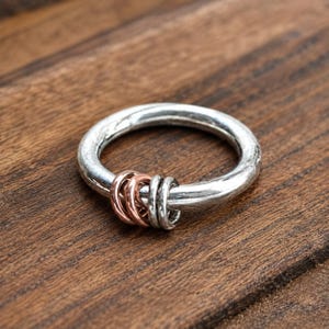May include: A silver-toned ring featuring a thick, textured band with four smaller, movable rings attached. Two of the smaller rings are copper-toned, while the other two are silver-toned, creating a contrasting metallic appearance on the band.