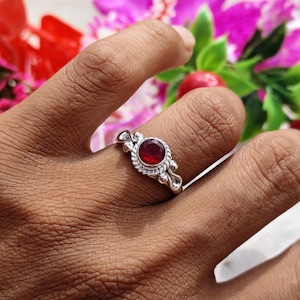 May include: A silver ring featuring a round, deep red gemstone set in a bezel. The metal band displays a twisted rope texture surrounding the stone, with small decorative silver spheres positioned at the top and bottom of the setting.