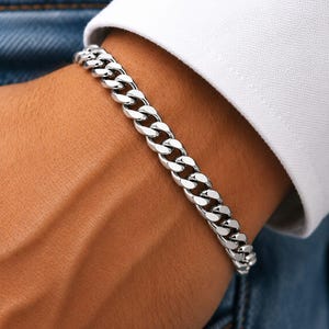 May include: A polished silver-tone curb chain bracelet featuring interlocking flat, rounded links. The bracelet has a high-shine metallic finish and is worn around the wrist, positioned against a white shirt cuff and blue denim fabric.