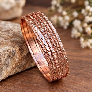May include: A set of five rose gold-toned metal bangles featuring a textured, diamond-cut surface pattern. Each bangle has a polished interior and a reflective, faceted exterior that catches the light, creating a shimmering effect across the set.