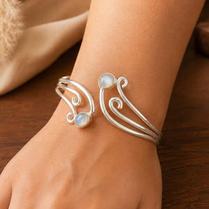 Könnte beinhalten: Ein silberfarbenes, offenes Armband mit einem mehrschichtigen Wirbeldesign an beiden Enden. Jede Seite des Armbands ist mit einem einzelnen, runden, schillernden Cabochon-Stein verziert, der einen sanften blauen Schimmer auf der metallischen Oberfläche zeigt.