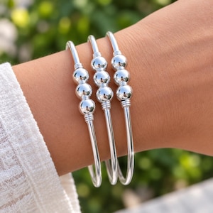 Könnte beinhalten: Drei polierte Armreifen in Silberoptik, die jeweils mit drei glatten, runden Perlen in der Mitte eines schmalen Metallbandes versehen sind. Die Armbänder werden zusammen getragen und zeigen eine reflektierende Oberfläche und ein Design.