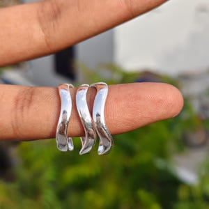 Op de afbeelding: Drie zilverkleurige ringen met een gladde, gepolijste afwerking en een opvallende, golvende, slangachtige vorm. Elke ring heeft een doorlopend gebogen ontwerp dat van opzij gezien een vloeiend, golvend silhouet creëert dat het licht reflecteert.