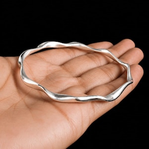 Pode incluir: Uma pulseira de prata polida com um design ondulado contínuo. A superfície metálica é lisa e reflexiva, formando uma forma circular com curvas suaves e rítmicas que criam uma aparência fluida ao redor de toda a pulseira.