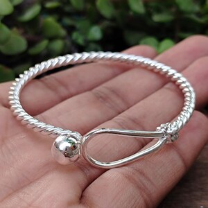 Puede incluir: Una pulsera de tono plateado con una banda trenzada similar a una cuerda. Un extremo de la pulsera termina en una cuenta esférica lisa y pulida, mientras que el otro extremo forma un cierre de gancho de bucle abierto y alargado.