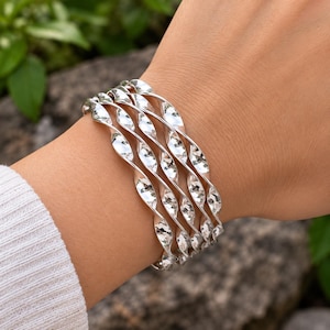 Puede incluir: Un conjunto de cuatro brazaletes plateados con un diseño de espiral retorcida continua. Cada brazalete tiene una superficie pulida y reflectante que capta la luz, creando una serie de segmentos metálicos curvos que se repiten alrededor.